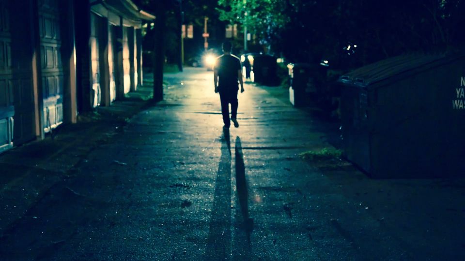 A lone figure walking down a dark, wet street at night under a streetlight.
