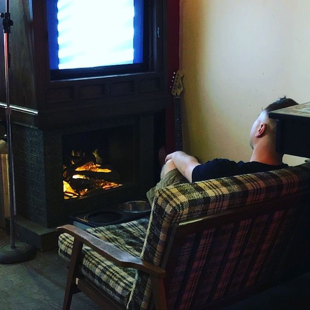 Seen from behind, a man relaxes in a plaid armchair beside a fireplace.