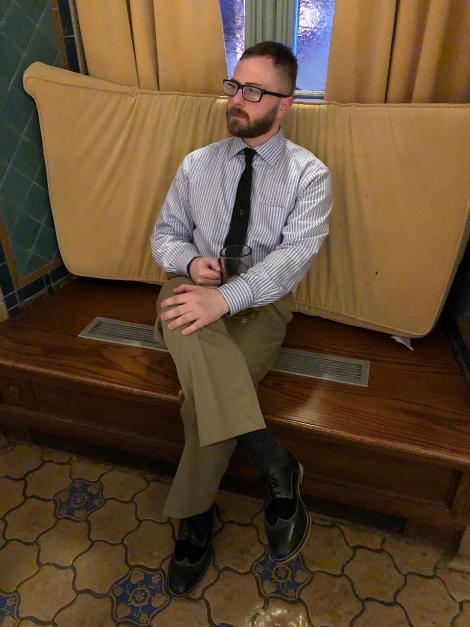 TakTrack in glasses, a striped shirt and tie, seated cross-legged on a wooden bench.