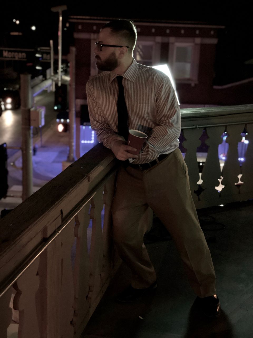 TakTrack in a dress shirt and tie leaning on a balcony railing above a city street at night.