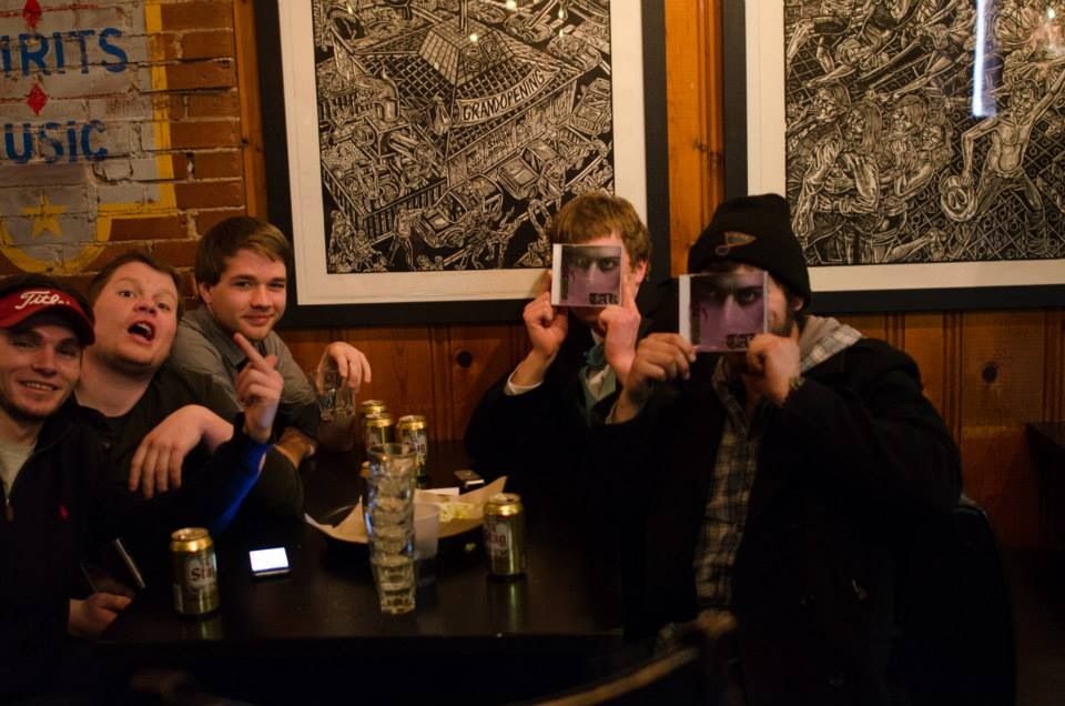 Friends crowded around a bar table with drinks, two of them holding photos over their eyes.
