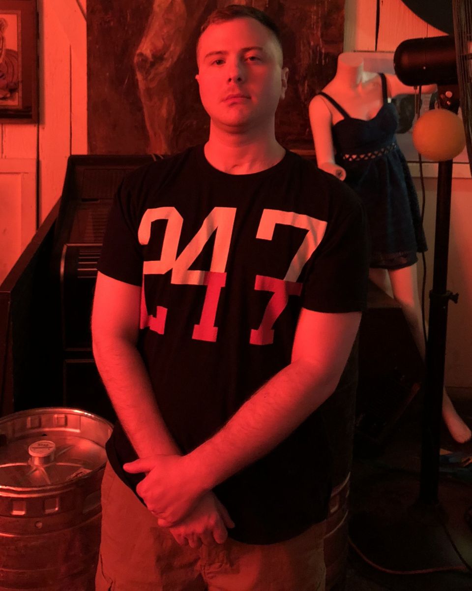TakTrack standing in a red-lit room, hands clasped in front of him, in a black T-shirt with large numbers.