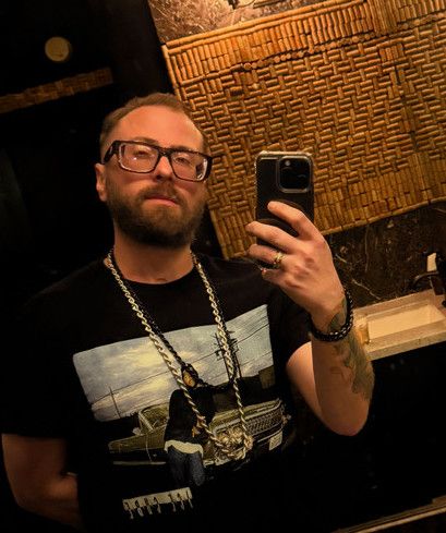 TakTrack taking a mirror selfie in glasses, a black graphic T-shirt and gold chains.