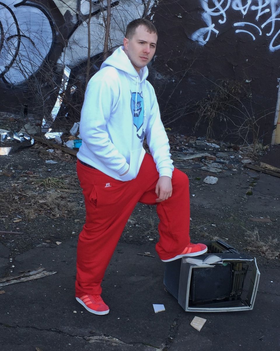 TakTrack in a white hoodie and red pants, one foot on a broken TV in a graffiti-covered lot.